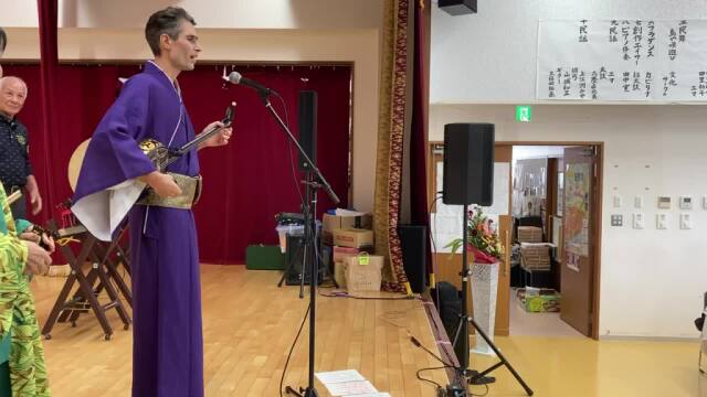Me standing on a stage in front of a micro, holding a sanshin
