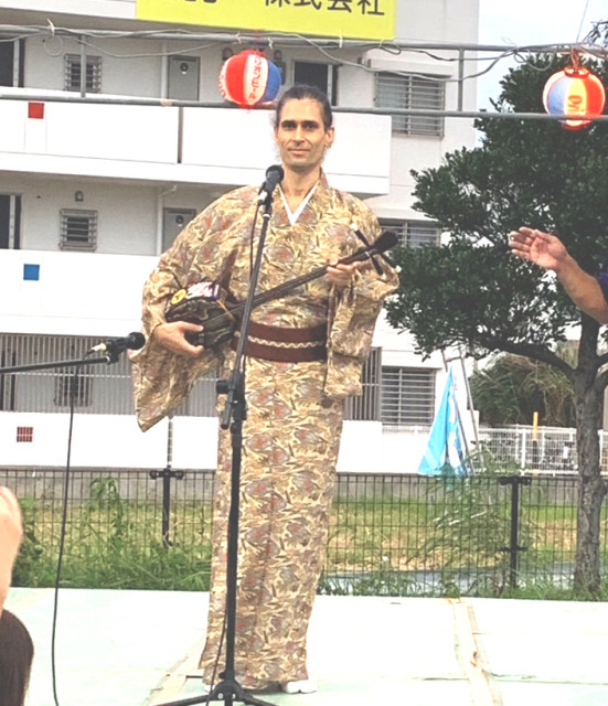 Me standing on a stage outdoors, holding a sanshin, in front of a microphone Me standing on a stage outdoors, holding a sanshin, in front of a microphone